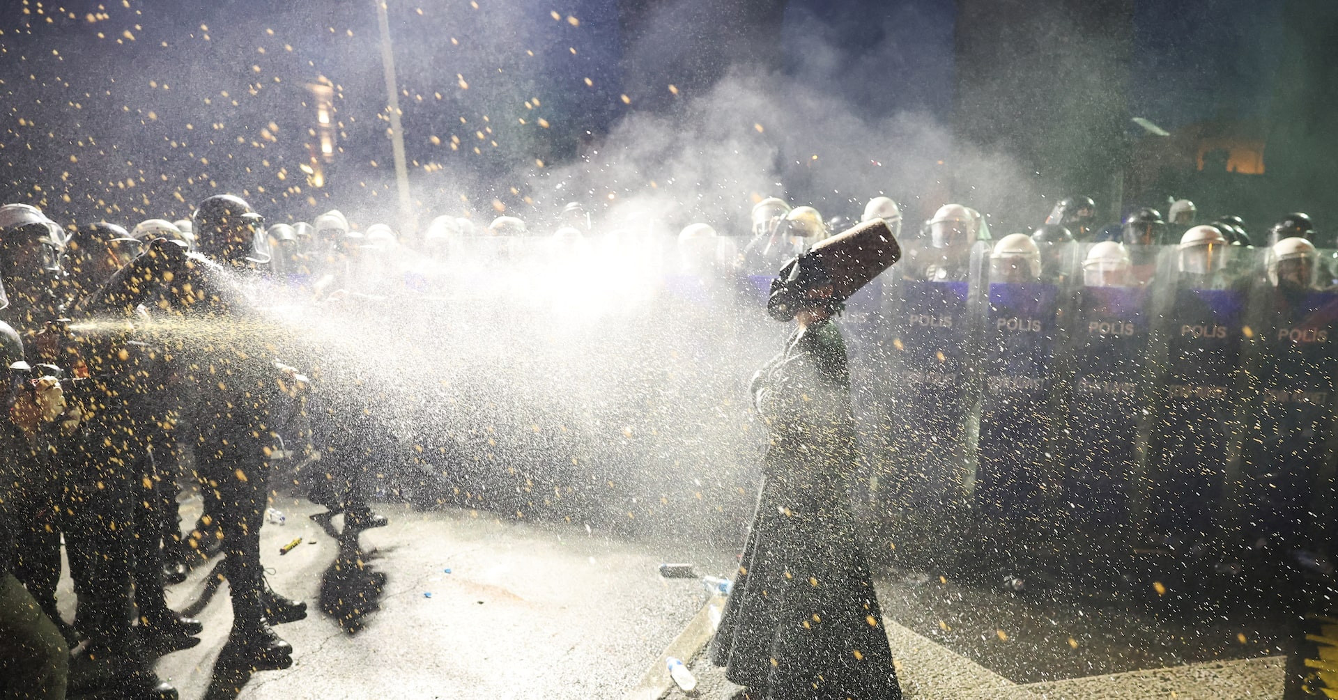 A person in a black robe and a tall brown felt hat stands facing a line of riot police holding shields labeled “Polis.” The person is being sprayed with pepper spray, and the air is filled with mist and debris. The person wears a gas mask and stands with a composed posture amid the chaos.